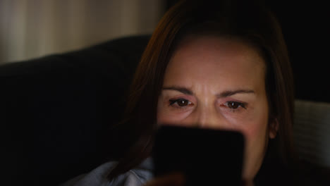 Close-Up-Of-Anxious-Woman-Sitting-On-Sofa-At-Home-At-Night-Looking-At-Mobile-Phone-Concerned-About-Social-Media-Or-Bad-News